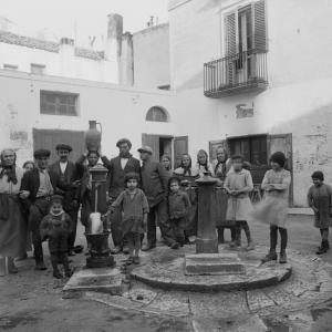Una fotografia storica in bianco e nero che ritrae un numeroso gruppo di abitanti di un paese riuniti attorno a delle fontane pubbliche nel cortile o piazzetta tra le case.
